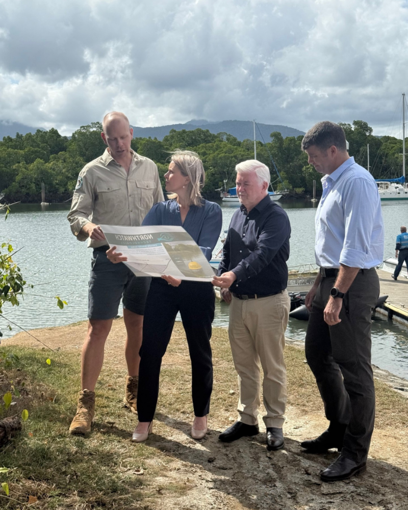 Photo shows two men and a woman looking at a map held by a man wearing shorts, a khaki shirt, and boots. They are outdoors, standing on a grassy area next to water with boats and docks visible in the background. 